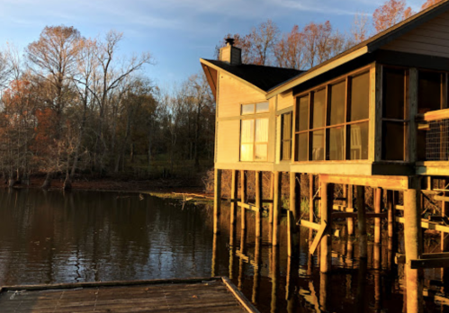 A lakeside house with large windows reflecting the sunset, surrounded by trees and calm water.