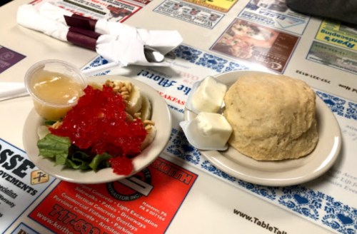 A plate with a biscuit, salad topped with red gelatin, and a small cup of applesauce on a patterned table.