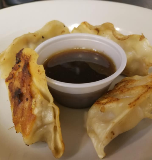A plate with five golden-brown dumplings arranged around a small cup of dark dipping sauce.
