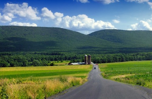 A winding road leads to a farm with silos, surrounded by lush green fields and rolling hills under a blue sky.