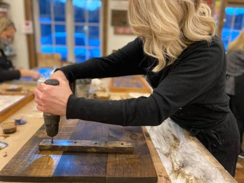 A person using a power drill on a wooden project in a workshop, with other crafting materials visible in the background.