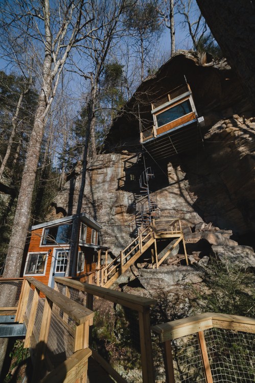 A unique treehouse built into a rock face, featuring wooden structures and a staircase surrounded by trees.
