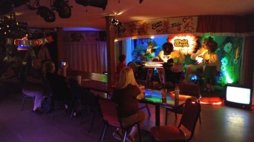 A dimly lit entertainment venue with a stage, colorful lights, and a small audience watching a performance.