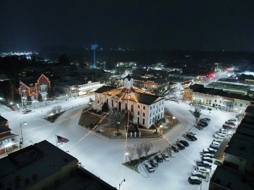 Aerial view of a snow-covered town square at night, featuring a historic building and festive lights.