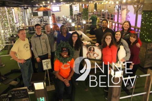 A group of people poses together in a lively indoor mini-golf setting, featuring fun decorations and vibrant lighting.