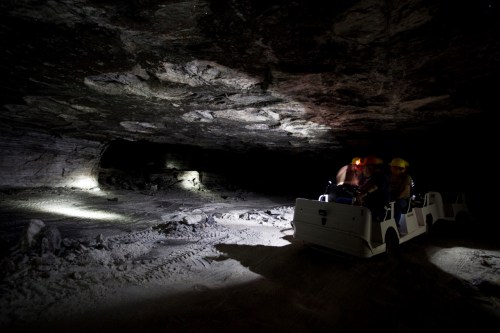 Two people in hard hats ride a small cart through a dimly lit underground mine with rocky walls.