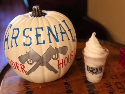 A white pumpkin painted with "Arsenal Cider House" and two crossed guns, next to a cup of soft serve ice cream.