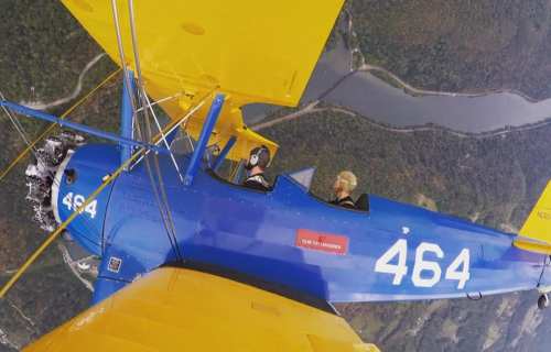 Aerial view of two pilots in a blue and yellow biplane flying over a river and green landscape.