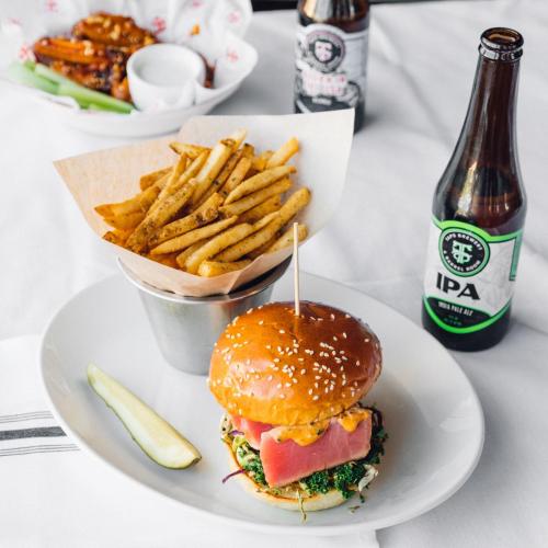 A sesame seed burger with greens, a pickle, crispy fries in a cup, and a bottle of IPA beer on a table.