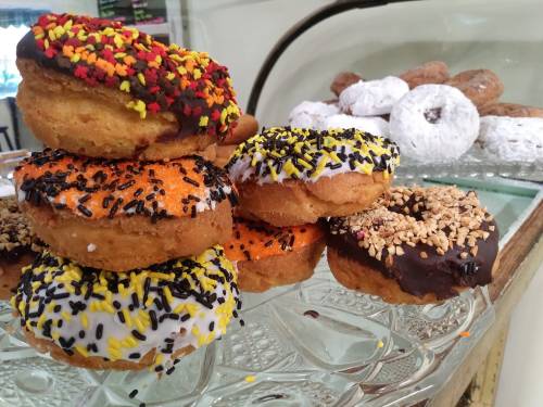 A display of colorful donuts topped with sprinkles, including chocolate, orange, and white icing, with powdered donuts in the background.