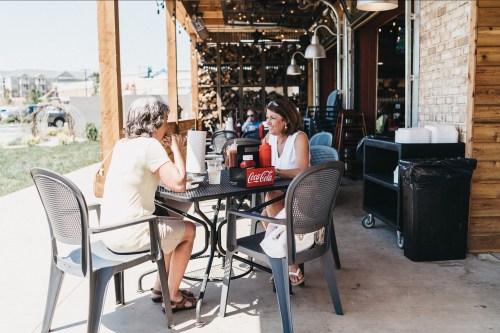 Two women sit at an outdoor table enjoying drinks and food at a casual restaurant.