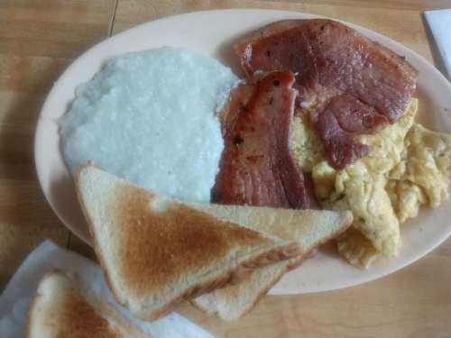A plate of breakfast featuring scrambled eggs, grits, bacon, and toasted bread.