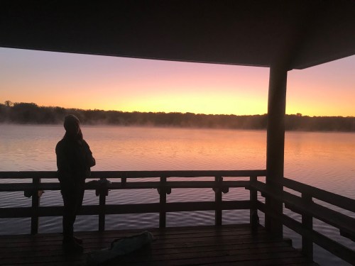A person stands on a dock, silhouetted against a colorful sunset over a calm lake with mist rising.