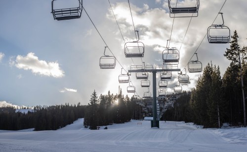 A ski lift towers over a snowy landscape, with trees and a cloudy sky in the background. Sunlight peeks through the clouds.