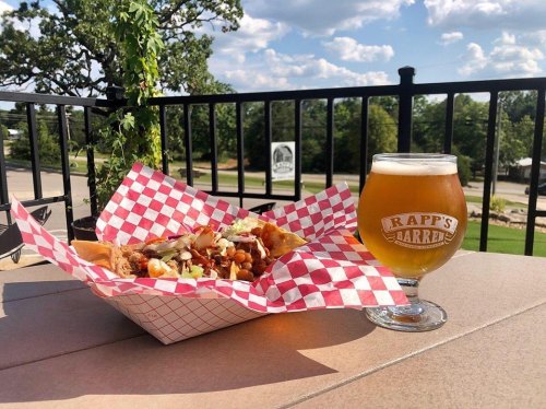 A plate of nachos with toppings beside a glass of beer on a table, with a scenic outdoor view in the background.