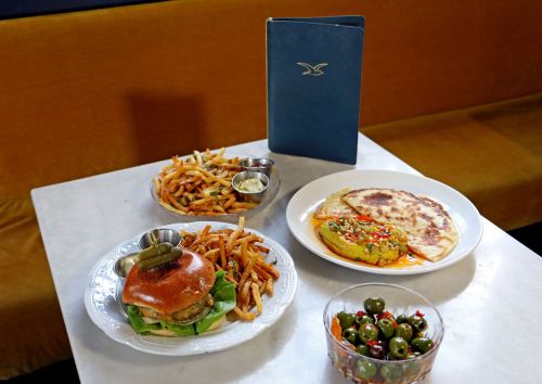 A table with a burger, fries, a flatbread dish, and a bowl of olives, alongside a menu on a blue cover.