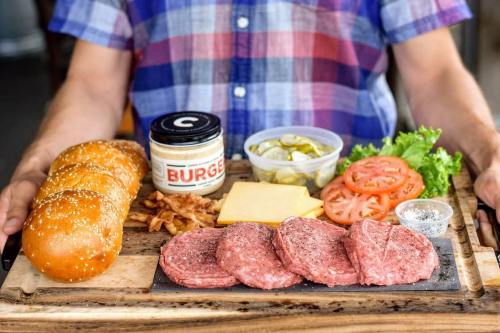 A person holds a wooden platter with burger ingredients: buns, patties, cheese, tomatoes, pickles, lettuce, and fries.
