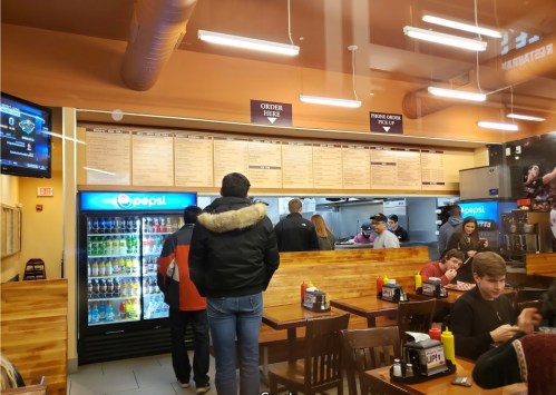 A busy restaurant interior with a menu board, customers ordering, and a drink fridge in the background.