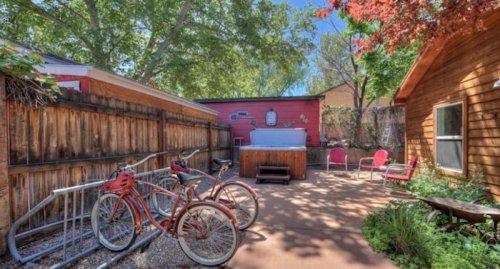 A sunny backyard with a hot tub, wooden fence, bicycles, and seating areas surrounded by trees.