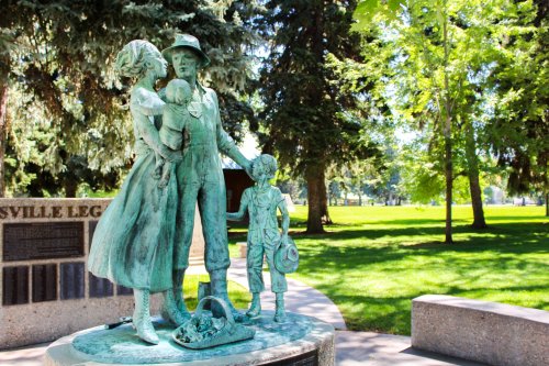 A bronze statue of a family, featuring a woman holding a child and a boy beside her, set in a green park.