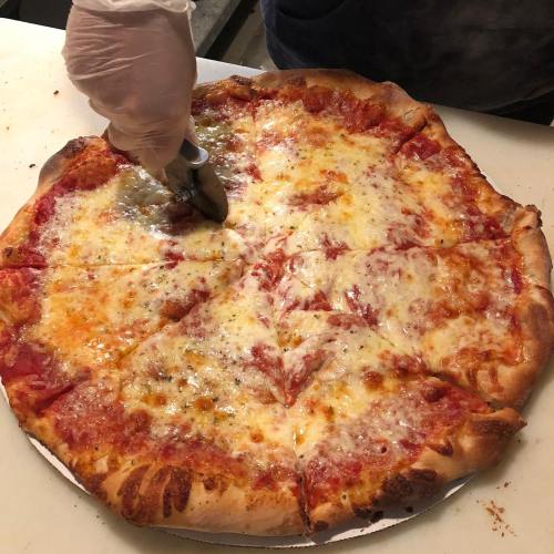A hand with a gloved finger uses a pizza cutter to slice a large cheese pizza on a white surface.