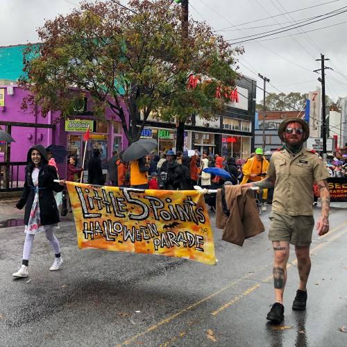 A festive Halloween parade with participants holding a colorful banner, walking in the rain.