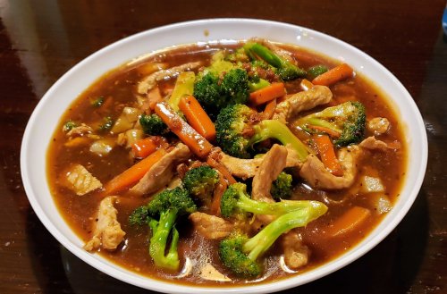 A bowl of stir-fried chicken with broccoli, carrots, and a savory brown sauce on a wooden table.