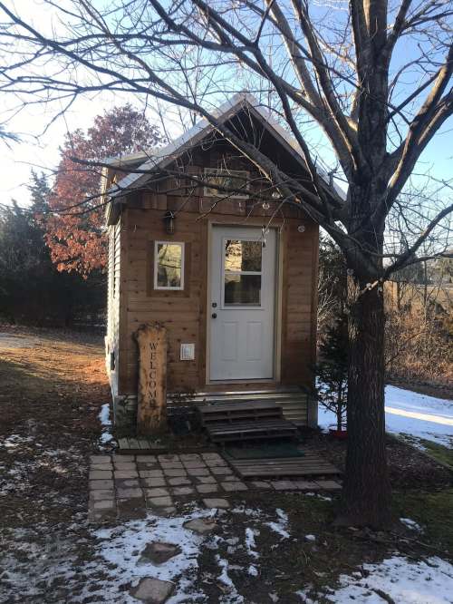 A small wooden cabin with a white door, surrounded by trees and snow, featuring a "Welcome" sign by the entrance.