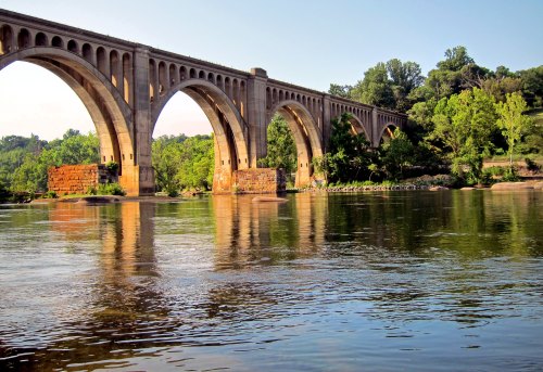 A large stone bridge with arches spans a calm river, surrounded by lush greenery and a clear blue sky.