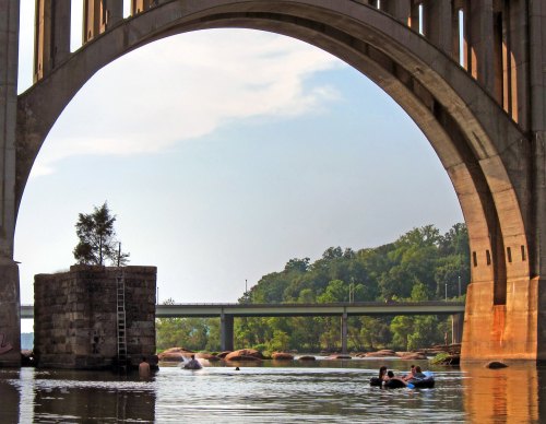 A serene river scene under a large arch bridge, with two people kayaking near a stone pillar and lush greenery in the background.