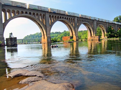 A large stone bridge with multiple arches spans a river, with a train on top and a small boat below.