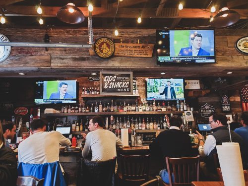 A lively bar scene with patrons seated at the counter, multiple TVs showing sports, and a rustic wooden decor.