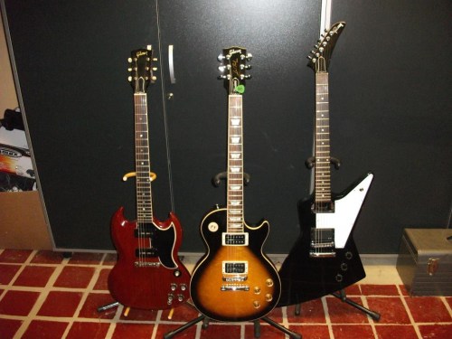 Three electric guitars on stands: a red SG, a sunburst Les Paul, and a black Explorer, set against a dark background.