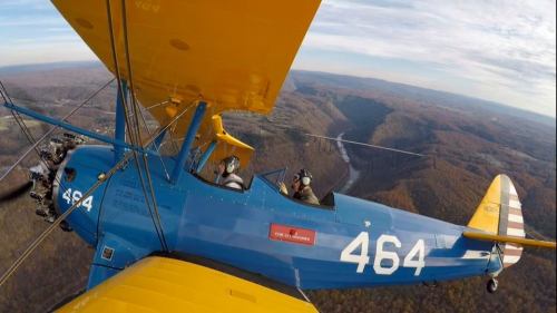 A blue biplane with yellow wings flying over a scenic landscape of hills and valleys under a cloudy sky.