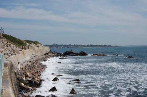A rocky shoreline with waves crashing against the rocks, under a clear blue sky and distant coastal town.