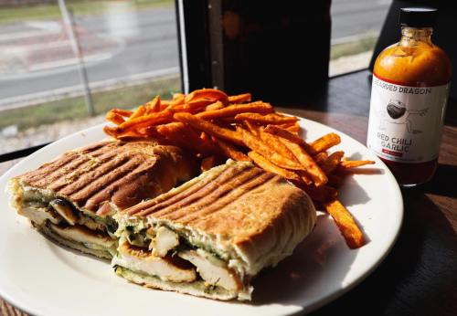 A plate with a grilled sandwich filled with chicken and greens, accompanied by sweet potato fries and a bottle of chili sauce.