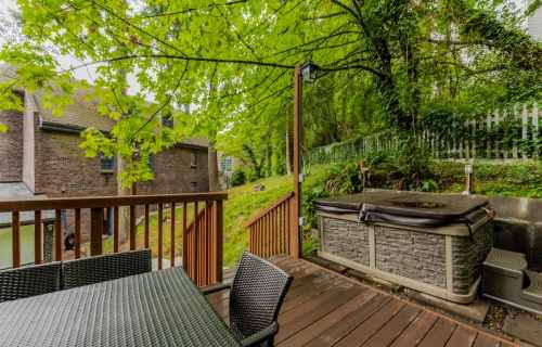 A wooden deck with a hot tub surrounded by lush greenery and a brick building in the background.