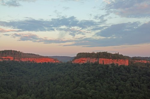 Sunset over rugged cliffs surrounded by lush green forests and a colorful sky with scattered clouds.
