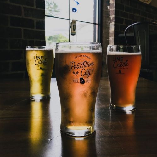 Three glasses of beer in varying colors on a wooden table, with a brick wall and window in the background.