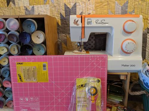 A sewing machine next to a cutting mat and rotary cutter, with colorful spools of thread in the background.