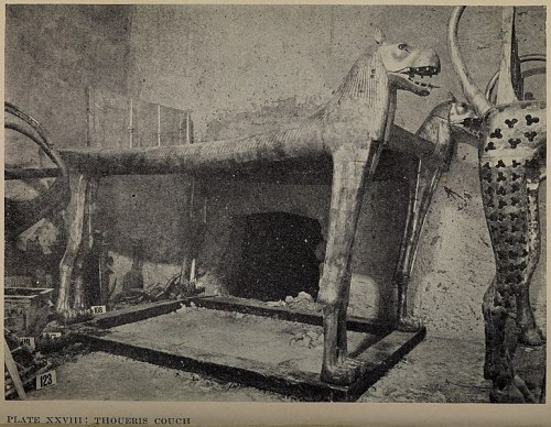 An ancient wooden couch shaped like a horse, displayed in a museum setting with other artifacts in the background.