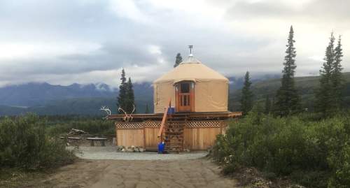 A cozy yurt on a wooden platform, surrounded by trees and mountains under a cloudy sky.