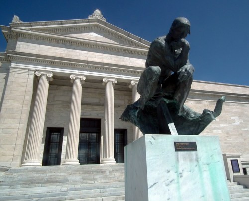 A bronze statue of a seated thinker in front of a grand stone building with columns and steps.