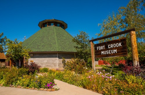 A round building with a green roof, surrounded by colorful flowers, featuring a sign that reads "Fort Crook Museum."