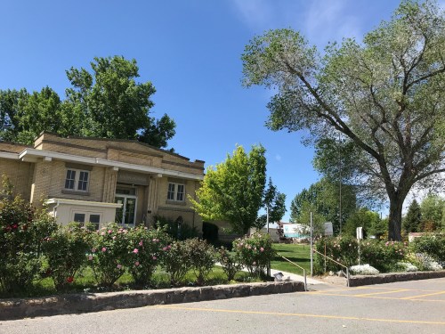A brick building surrounded by green trees and blooming rose bushes under a clear blue sky.