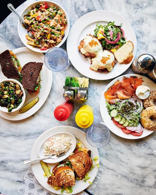 A spread of colorful dishes on a marble table, including salads, sandwiches, bagels, and sides.