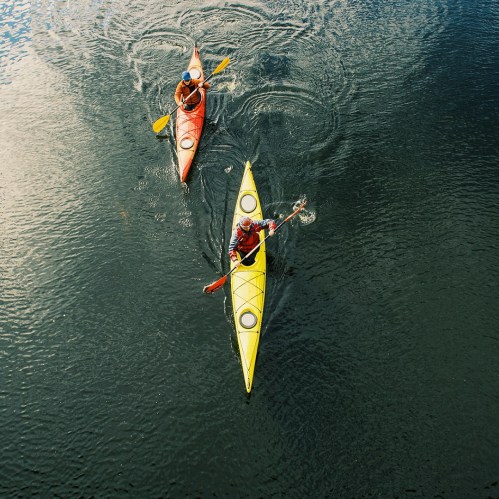 Two kayakers paddle on a calm water surface, one in a yellow kayak and the other in red.
