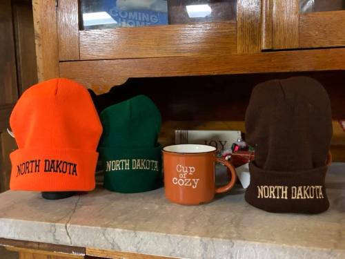 Three beanie hats in orange, green, and brown with "NORTH DAKOTA" embroidered, alongside a mug that says "Cup of Cozy."
