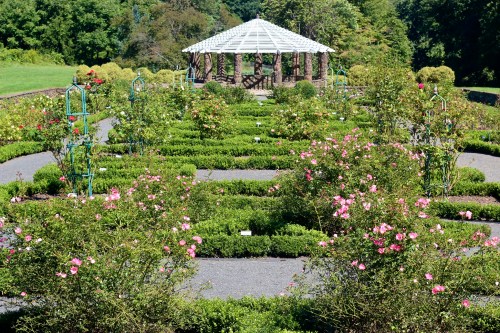 A landscaped garden featuring neatly arranged flower beds, a gazebo, and lush greenery under a clear blue sky.