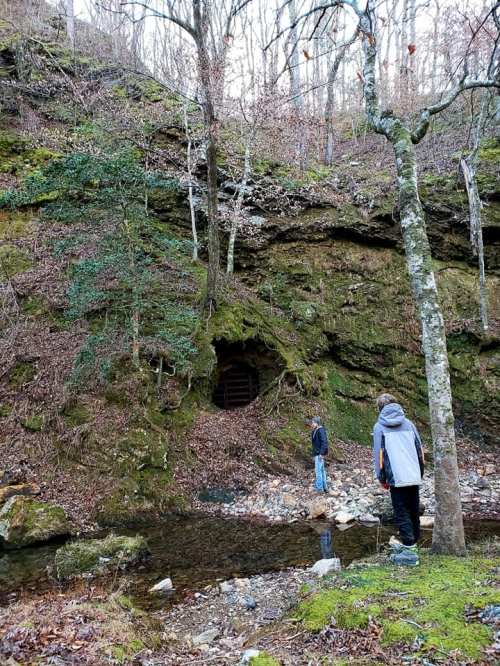Two people stand near a mossy rock formation with a small cave entrance, beside a shallow stream in a wooded area.